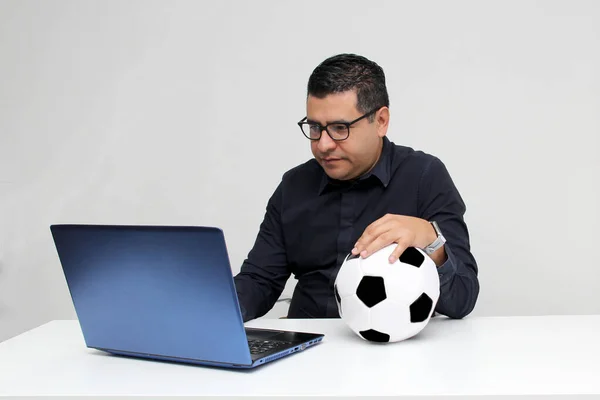 stock image Latino adult office man watches football games on his work laptop during office hours in the morning, he sees him nervous, sad, stressed, angry next to his soccer ball