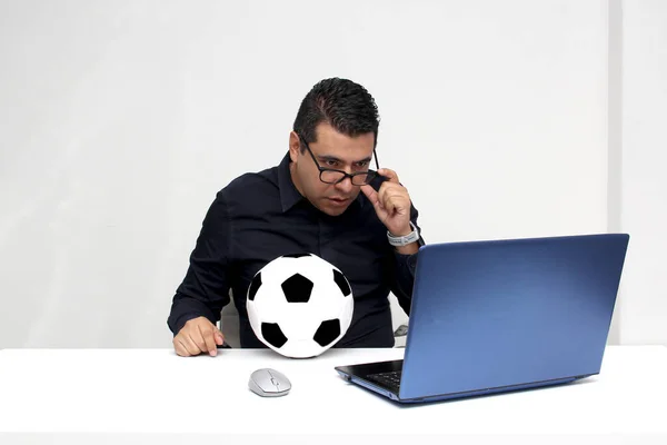 Latino adult office man watches football games on his work laptop during office hours in the morning, he sees him nervous, sad, stressed, angry next to his soccer ball