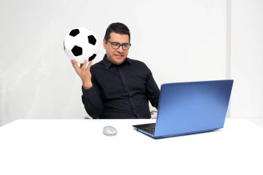 Latin yetişkin ofis çalışanı, iş saatleri boyunca futbol maçlarını bilgisayarından izliyor. Onu futbol topunun yanında heyecanlı, gergin ve şaşırmış görüyor.