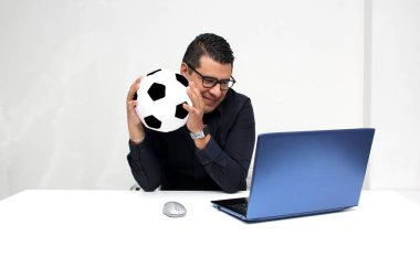Latin yetişkin ofis çalışanı, iş saatleri boyunca futbol maçlarını bilgisayarından izliyor. Onu futbol topunun yanında heyecanlı, gergin ve şaşırmış görüyor.