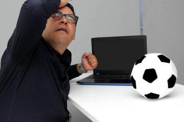 Latin yetişkin ofis çalışanı, iş saatleri boyunca dizüstü bilgisayarından futbol maçlarını izliyor. Onu futbol topunun yanında sinirli, üzgün, stresli ve sinirli görüyor.
