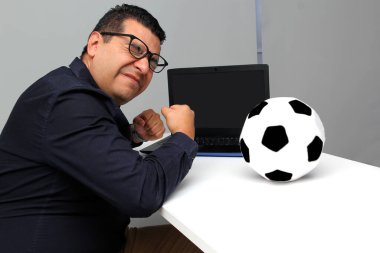 Latin yetişkin ofis çalışanı, iş saatleri boyunca dizüstü bilgisayarından futbol maçlarını izliyor. Onu futbol topunun yanında sinirli, üzgün, stresli ve sinirli görüyor.