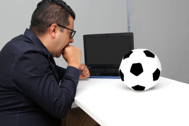Latin yetişkin ofis çalışanı, iş saatleri boyunca dizüstü bilgisayarından futbol maçlarını izliyor. Onu futbol topunun yanında sinirli, üzgün, stresli ve sinirli görüyor.