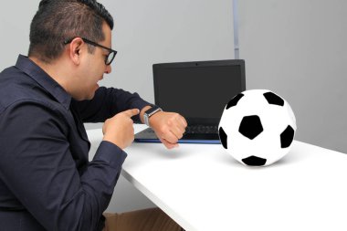 Latin yetişkin ofis çalışanı, iş saatleri boyunca dizüstü bilgisayarından futbol maçlarını izliyor. Onu futbol topunun yanında sinirli, üzgün, stresli ve sinirli görüyor.