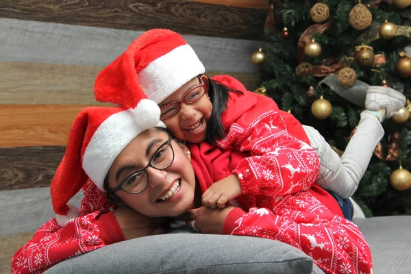 Single divorced mom and daughter Latina brunettes with glasses dressed in pajamas play in bed next to the Christmas tree with Santa hats give each other hugs and kisses show their love in solitude