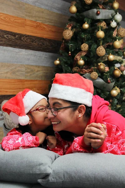 Single divorced mom and daughter Latina brunettes with glasses dressed in pajamas play in bed next to the Christmas tree with Santa hats give each other hugs and kisses show their love in solitude