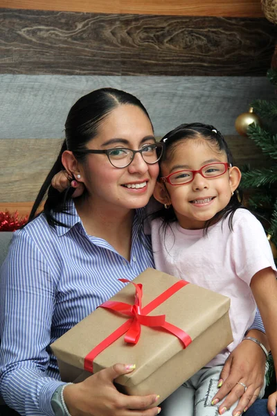 Divorced single mom and daughter Latina brunettes with glasses have Christmas presents sitting by the tree with spheres give each other hugs and kisses show their love in solitude