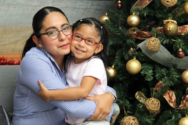 Divorced single mom and daughter Latina brunettes with glasses have Christmas presents sitting by the tree with spheres give each other hugs and kisses show their love in solitude