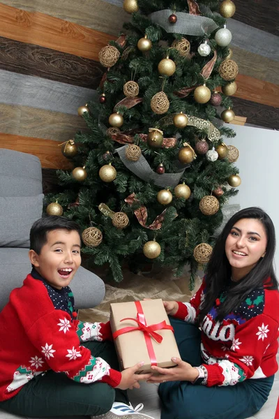 Divorced single mom and son Latino have Christmas presents sitting by the tree with spheres give each other hugs and kisses show their love in solitude