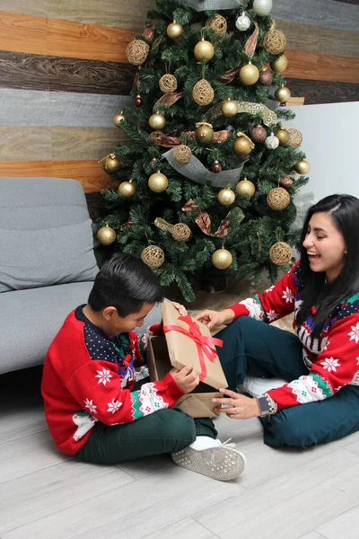 Divorced single mom and son Latino have Christmas presents sitting by the tree with spheres give each other hugs and kisses show their love in solitude