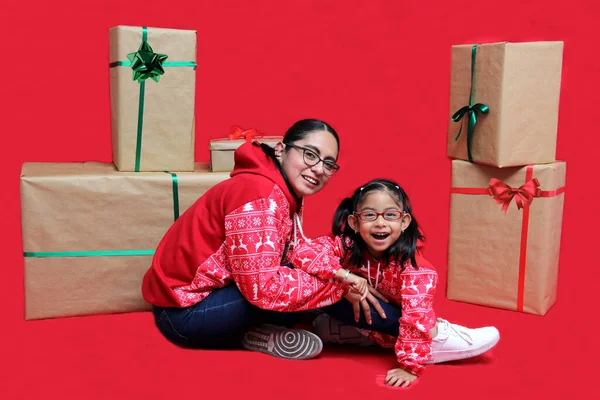 Latina mom and daughter with glasses wear ugly Christmas sweaters and show their love to each other on a red background between large gift boxes with bow