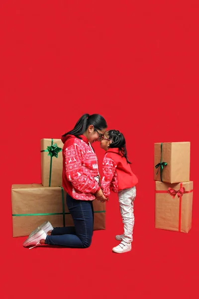 Latina mom and daughter with glasses wear ugly Christmas sweaters and show their love to each other on a red background between large gift boxes with bow