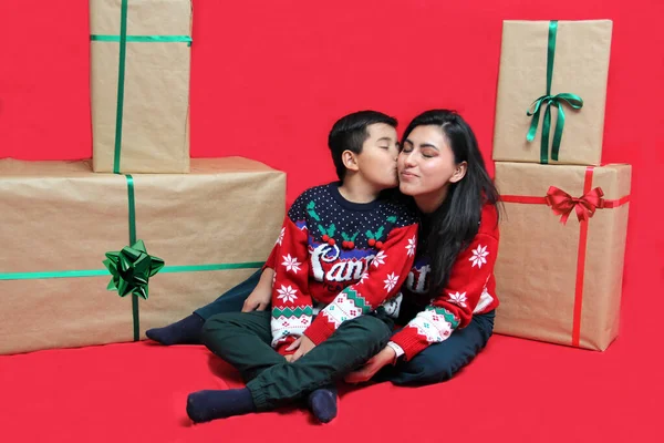 Latina mom and son wear ugly Christmas sweaters and show their love to each other on a red background between large gift boxes with bow