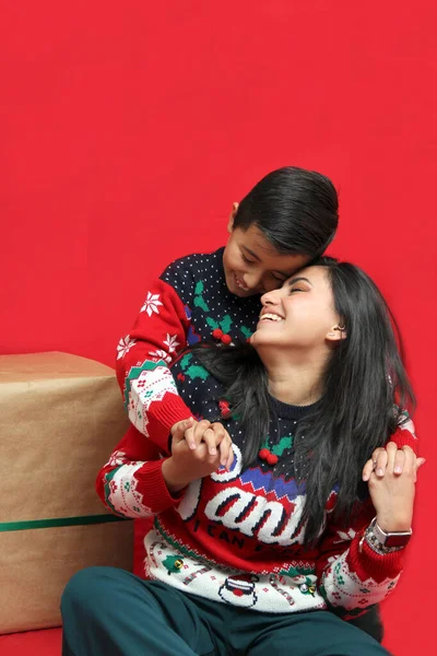 Latina mom and son wear ugly Christmas sweaters and show their love to each other on a red background between large gift boxes with bow