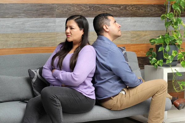 Latino adult couple of man and woman sitting on a sofa are upset and angry about problems that may cause their separation
