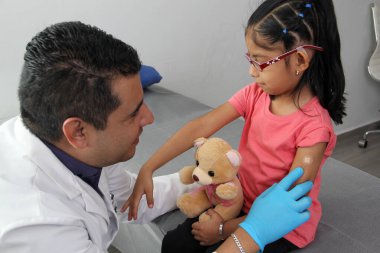 Dark-haired Latino doctor and little girl have a medical consultation in the pediatric office to vaccinate their arm against Covid, chickenpox, diphtheria, influenza, hepatitis, measles, mumps clipart