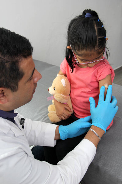 Dark-haired Latino doctor and little girl have a medical consultation in the pediatric office to vaccinate their arm against Covid, chickenpox, diphtheria, influenza, hepatitis, measles, mumps