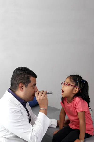 Professional specialist pediatrician doctor and patient 4-year-old Latino brown-haired girl are in consultation to check their eyes, mouth and throat to diagnose any disease in the office