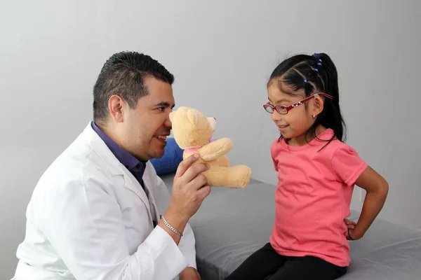Latino pediatric specialist doctor plays with his patient a 4-year-old girl relaxes her with a teddy bear as a toy to reassure her in her office