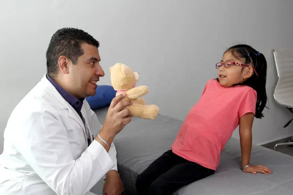 Latino pediatric specialist doctor plays with his patient a 4-year-old girl relaxes her with a teddy bear as a toy to reassure her in her office