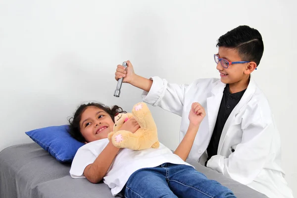 Latino girl and boy play to be a doctor with a white coat that is too big for her and patient who acts to feel sick very entertaining and funny they make jokes
