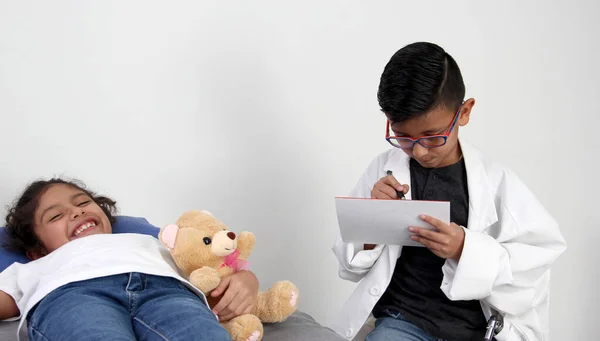 Latino girl and boy play to be a doctor with a white coat that is too big for her and patient who acts to feel sick very entertaining and funny they make jokes