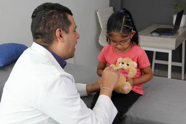Latino pediatrician doctor checks his girl patient with autism spectrum disorder ASD, he communicates through his teddy bear that he carries as an attachment or transitional object