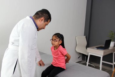 Latino pediatrician doctor attends to his 4-year-old brunette girl patient with headache, migraine and dizziness in his office wearing a white coat