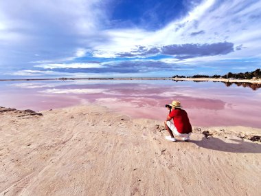 Şortlu, kırmızı tişörtlü ve şapkalı Latin yetişkin fotoğrafçı Yucatan Meksika 'da tuz yoğunluğu yüksek olan pembe lagünün yanındaki kumda fotoğraf çekiyor.