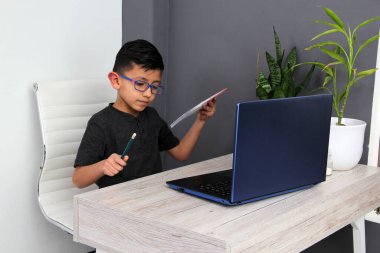 7-year-old Latino boy with glasses does home schooling takes online classes at home on a desk with a laptop, studies, is surprised and participates in class