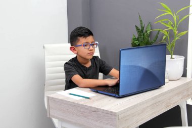 7-year-old Latino boy with glasses does home schooling takes online classes at home on a desk with a laptop, studies, is surprised and participates in class