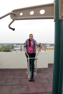 Yetişkin 40 yaşındaki Latin kadın, apartmanının çatı bahçesinde ortak egzersiz ekipmanıyla egzersiz yapıyor.