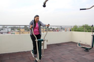 Yetişkin 40 yaşındaki Latin kadın, apartmanının çatı bahçesinde ortak egzersiz ekipmanıyla egzersiz yapıyor.