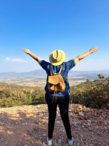 stock image Adult woman with hat looks towards the horizon on top of a mountain opens his arms and breathes in front of the mountainous landscape proud of her achievement