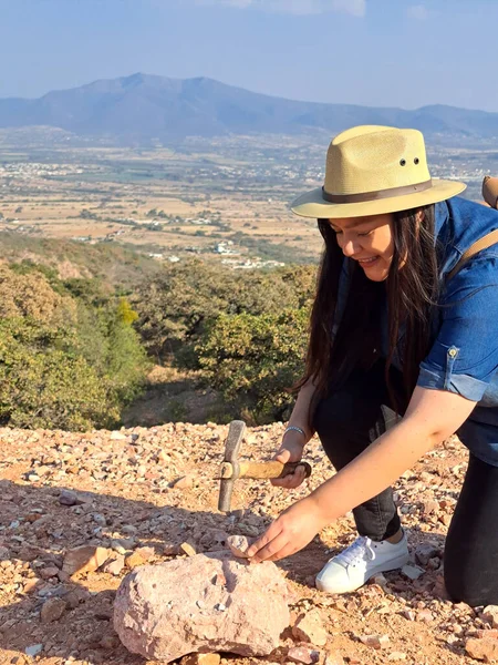 Latina woman with hat and miner's pick hammer works as a geologist, studies the composition and structure of the mineral soil of the mountain