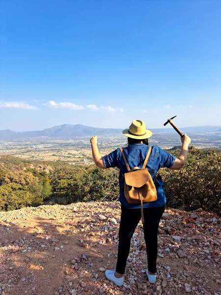 Latina woman with hat and miner\'s pick hammer works as a geologist, studies the composition and structure of the mineral soil of the mountain