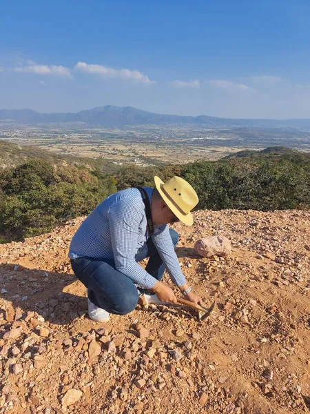 Latin man with hat and miner\'s pick hammer works as a geologist, studies the composition and structure of the mineral soil of the mountain