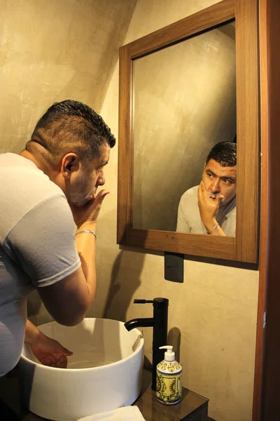 40歳の暗い肌のラテン系の大人の男は 彼がワックスを恐れているので 自宅でバスルームミラーの前にカミソリで剃刀で剃る — ストック写真