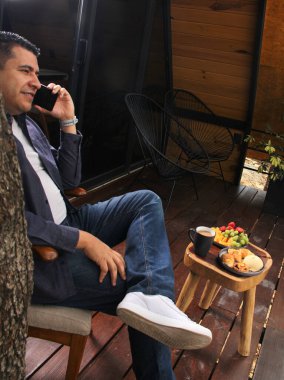 Siyah saçlı, 40 yaşındaki Latin yetişkin bir adam, ormanın ortasında bir kulübede kahvaltı ederken video ve mesajlar için cep telefonunu kullanıyor.