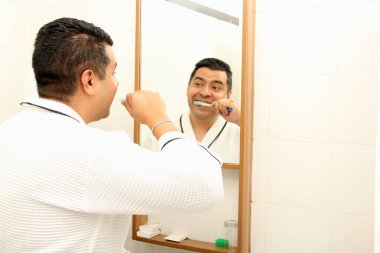 Kırk yaşında esmer tenli Latin bir adam banyoda bornozla dişlerini fırçalar aynaya bakar ve oral sağlığıyla ilgilenir.