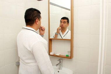 Kırk yaşında esmer tenli Latin bir adam banyoda bornozla dişlerini fırçalar aynaya bakar ve oral sağlığıyla ilgilenir.