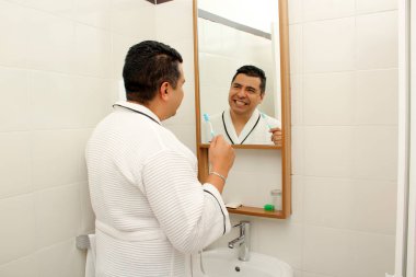 Kırk yaşında esmer tenli Latin bir adam banyoda bornozla dişlerini fırçalar aynaya bakar ve oral sağlığıyla ilgilenir.