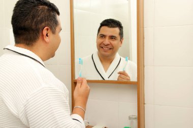 Kırk yaşında esmer tenli Latin bir adam banyoda bornozla dişlerini fırçalar aynaya bakar ve oral sağlığıyla ilgilenir.