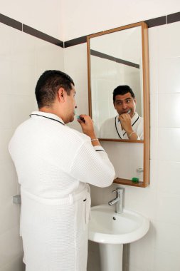 Kırk yaşında esmer tenli Latin bir adam banyoda bornozla dişlerini fırçalar aynaya bakar ve oral sağlığıyla ilgilenir.