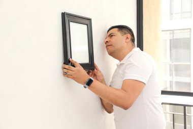 Koyu renk saçlı, 40 yaşında Latin bir adam yeni dairesini dekore etmiş. Bir emlakçıdan satın almış. Duvara bir çerçeve asmış.