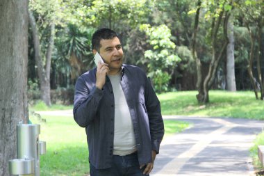 Parktaki 40 yaşındaki koyu renk saçlı Latin adam cep telefonunu uzaktan çalışmak için kullanıyor, telefon ediyor, randevu alıyor, internette alışveriş yapıyor.