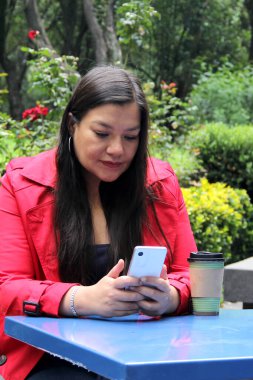 40 yaşında Latin bir kadın cep telefonuyla video görüşmesi yapıyor, internetten alışveriş yapıyor, randevularını alıyor ve açık hava bankında ödeme yapıyor.