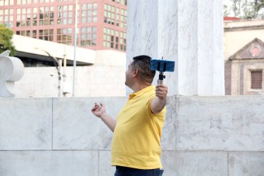 Siyah saçlı, yetişkin bir adam cep telefonuyla video kaydediyor. İnternetten canlı yayın yapıyor. Sokakta etkili biri tavsiyelerde bulunuyor.