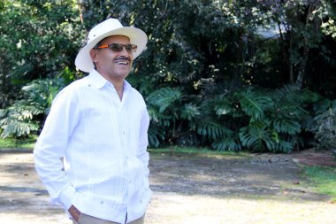 60 'larında yaşlı esmer tenli Latin bir adam emekliliğinde guayabera, şapka ve güneş gözlüğü giyerek parkta rahatlamış görünüyor.