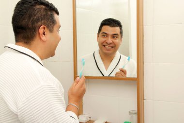 Esmer tenli 40 yaşındaki Latin adam diş dökülmesini ve çürükleri önlemek için banyo aynasının önünde dişlerini fırçalıyor.